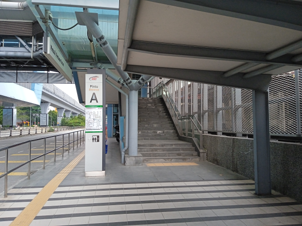 Ein Bahnsteig mit Treppen, eine Straße mit Geländern auf der linken Seite und ein Hintergrund mit Bäumen, Pfählen, Gebäuden und einer Brücke; der Bahnsteig hat Rohre und eine Tafel oben.