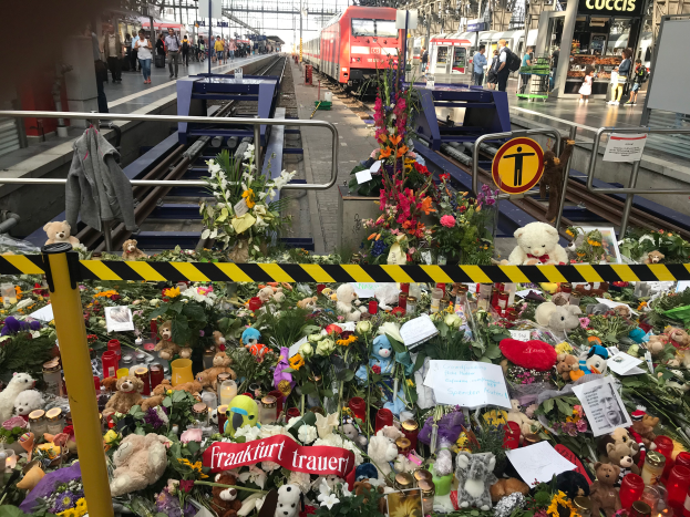 Ein Bahnsteig mit einem Zug im Hintergrund, umgeben von Menschen und verschiedenen Gegenständen wie Teddybären, Blumenbouquets und Kerzen, die an einen tragischen Zugunglück in Berlin, Deutschland, erinnern.