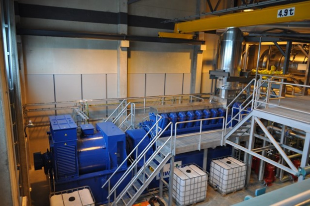 Ein Fabrikinnenraum mit Tr├Ągern, einer Treppe, Gel├Ąnder, Containern, L├Ąmpchen, Rohren und W├Ąnden.