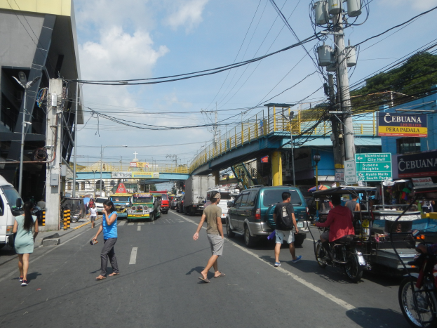 Eine belebte Stadtstraße mit Autos, Motorrädern, Fußgängern, Gebäuden, Strommasten, Laternen, Schildern, einer Brücke, Bäumen und einem bewölkten Himmel.