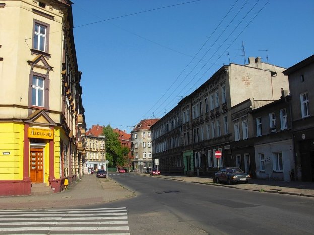 Stadtstraße mit fahrenden Autos, einer Zebrastreifen, Gebäuden auf beiden Seiten, Schildern auf Pfählen, Bäumen und einem klaren blauen Himmel.