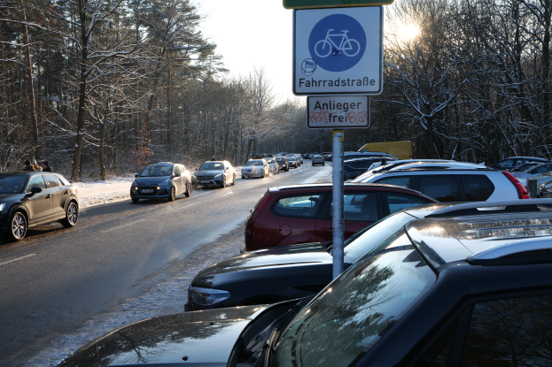 Ein Stau auf einer verschneiten Straße mit parkenden Autos am Straßenrand, ein Schild an einem Pfahl im Vordergrund und Bäume mit einem klaren Himmel im Hintergrund.