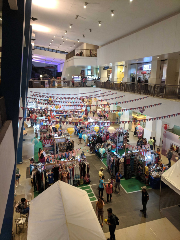 Ein volles Fest-Einkaufszentrum mit Ständen, hängenden Kleidern, Dekorationsfahnen, Ballons, sitzenden und stehenden Menschen, Geländern, Säulen und Deckenleuchten, ähnlich einem Weihnachtsmarkt im Mall of Asia.