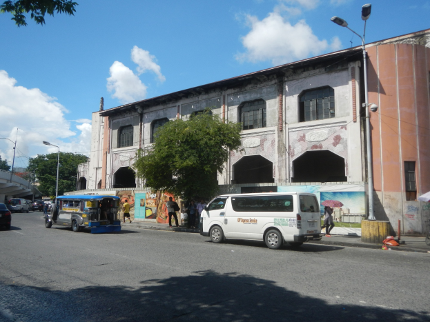 Eine belebte Stadtstraße mit Autos, Fußgängern, Bäumen, Laternen, einem großen Gebäude mit Wandgemälde, einer fernen Brücke und einem bewölkten Himmel.