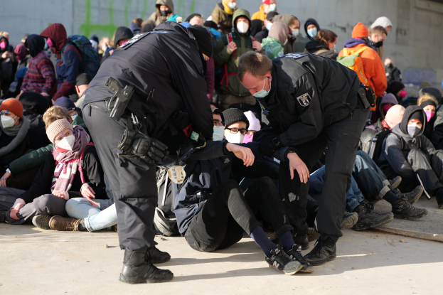 Eine Gruppe von Menschen, einige mit Mützen und Masken, sitzt auf dem Boden neben zwei Polizeibeamten, vor einer Wand mit Schrift, die auf eine städtische Umgebung hindeutet.