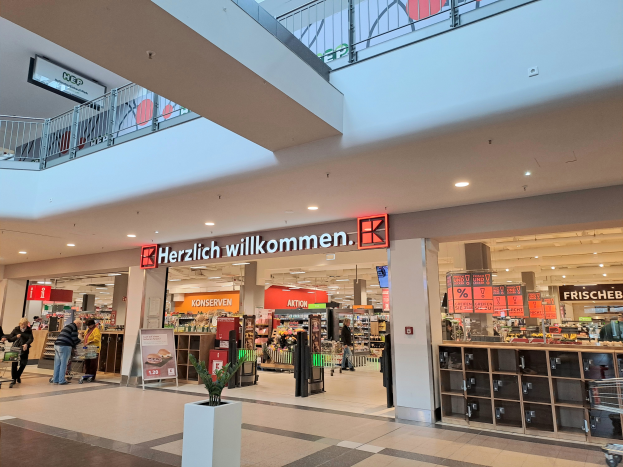 Das Innere eines Einkaufszentrums mit Menschen, die herumlaufen, Geschäften, Schildern mit Text, Lichtern, Geländern und einer Topfpflanze unten; der Text auf den Schildern lautet "Herzlich willkommen".
