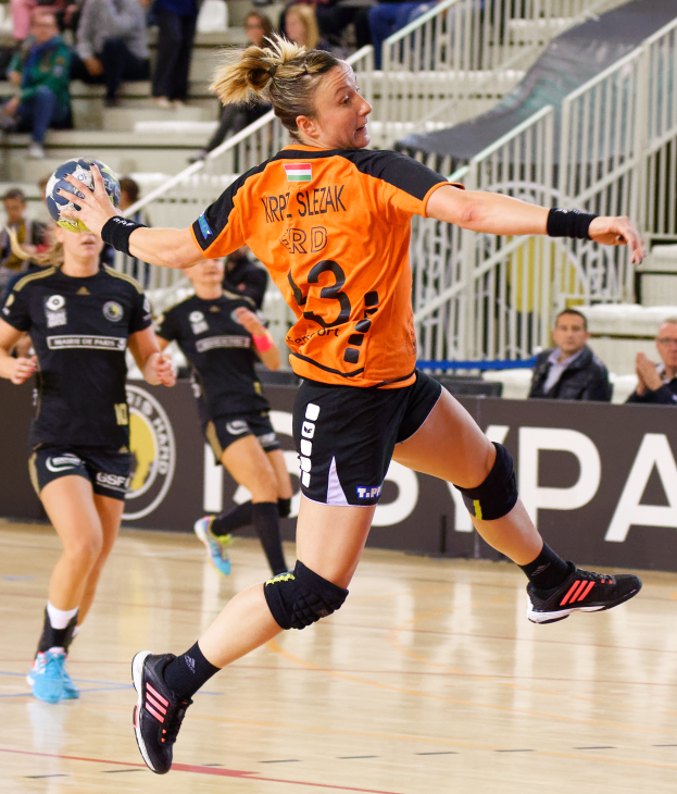 Eine Frau in einem orangen T-Shirt und schwarzen Shorts, die Handball spielt und einen Ball hält, mit Zuschauern und einer Tafel mit der Aufschrift "Krp Slezak - Handball-Weltmeisterschaft 2019" im Hintergrund.