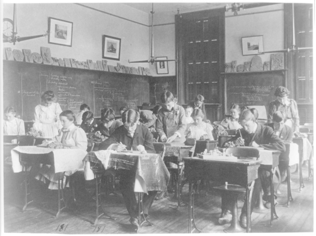 Ein Schwarz-Weiß-Foto eines Klassenzimmers mit Kindern, die an Tischen schreiben, verschiedenen Gegenständen auf den Tischen, Fotorahmen und einer Uhr an den Wänden und einer Tür im Hintergrund.
