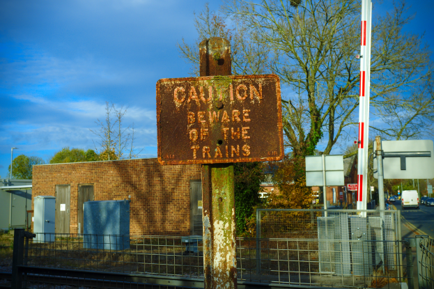 Eine Warnschilder an der Seite einer Bahnschiene, umgeben von einem Metallzaun, mit Bäumen, Pfählen, einem Gebäude mit Fenstern, Containern, einem Straßenpfahl, Fahrzeugen auf der Straße und einem bewölkten Himmel.