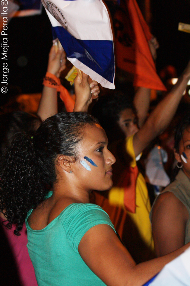 Eine Frau in einem grünen T-Shirt steht bei einer Demonstration und hält ein weißes Schild in der Hand, hinter ihr stehen viele andere Menschen.