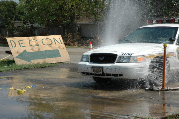 Ein Streifenwagen fährt durch eine Pfütze, mit einem Schild, Verkehrskegel und anderen Gegenständen auf dem Boden, mit Bäumen, Gebäuden und einem klaren blauen Himmel im Hintergrund.