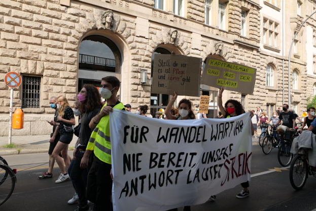 Eine Gruppe von Menschen marschiert auf einer Straße bei einer Demonstration, einige halten Schilder und fahren Fahrräder, im Hintergrund ein Gebäude mit Fenstern, Bögen, Säulen und Skulpturen.