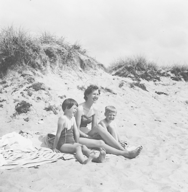 Schwarz-weiß-Foto einer Frau in einem Bikini und zweier Kinder in Badebekleidung, die auf einer Decke auf einem sandigen Strand mit Gras und Himmel im Hintergrund sitzen.