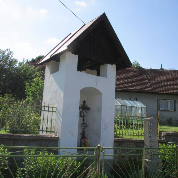 Kleines weißes Gebäude mit einem Kreuz darauf, umgeben von einem Metallzaun, gelegen in einem grasigen Bereich mit Pflanzen, einem Weg, nahen Häusern, Pfählen, Drähten und einem bewölkten Himmel.