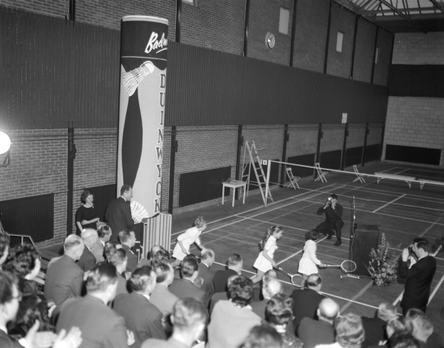 Ein Schwarz-Weiß-Foto einer Gruppe von Menschen, die Tennis in einer Turnhalle spielen, wobei einige stehen und andere auf Stühlen sitzen und Schläger halten, und eine Wand mit einer Uhr im Hintergrund.