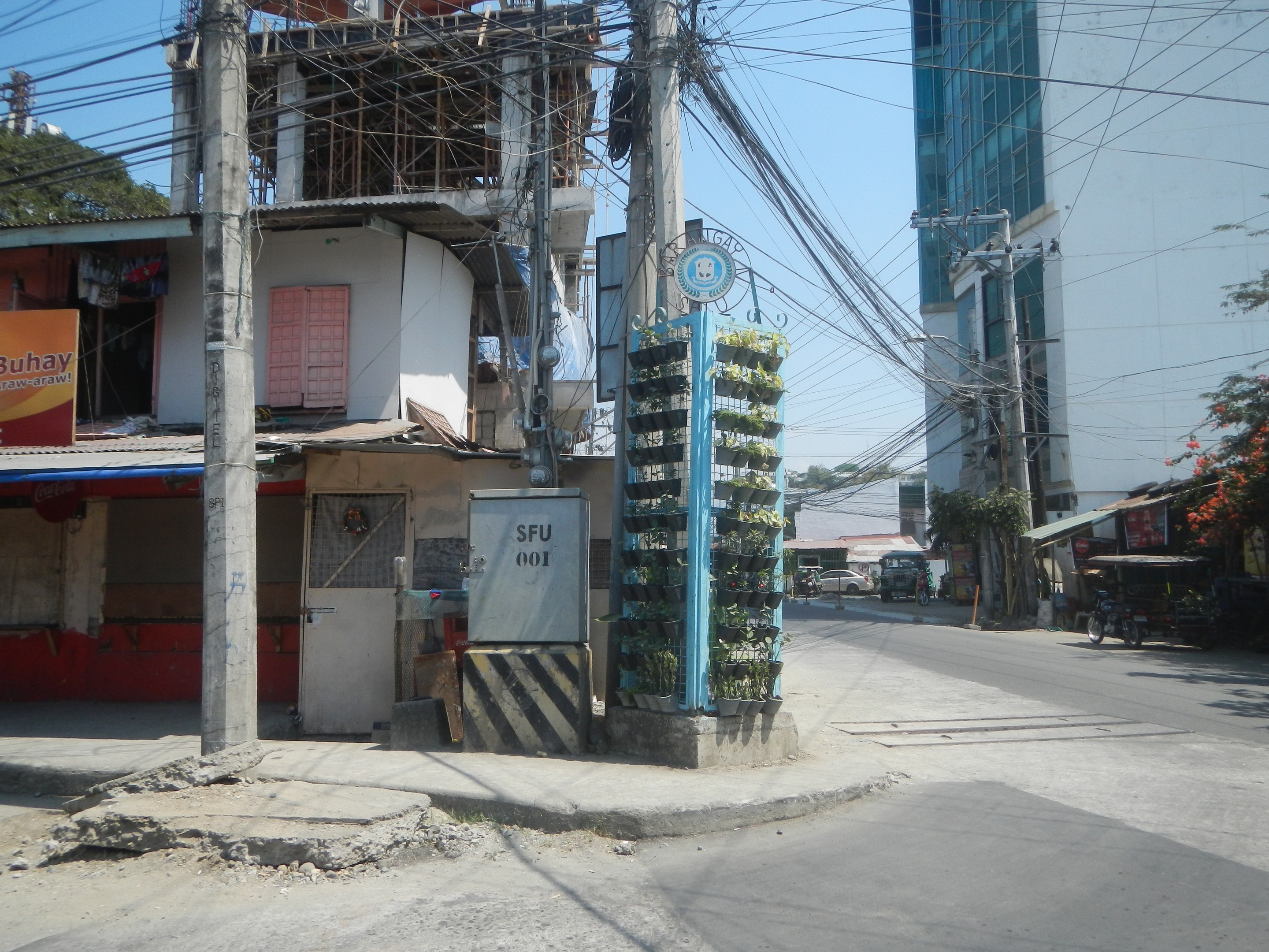 Stadtstraße mit Gebäuden, Strommasten, Kabeln, Schildern, Fahrzeugen, Bäumen und einem Gebäude mit Drähten im Vordergrund unter einem Himmel.