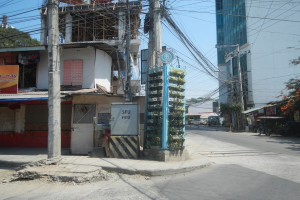 Stadtstraße mit Gebäuden, Strommasten, Kabeln, Schildern, Fahrzeugen, Bäumen und einem Gebäude mit Drähten im Vordergrund unter einem Himmel.