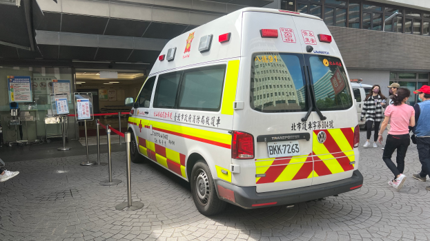 Ein Rettungswagen steht vor einem Gebäude mit Menschen auf der rechten Seite, eine Person sitzt auf einem Stuhl im Hintergrund und Schilder auf der linken Seite mit der Aufschrift "Rettungsdienst in Hong Kong".