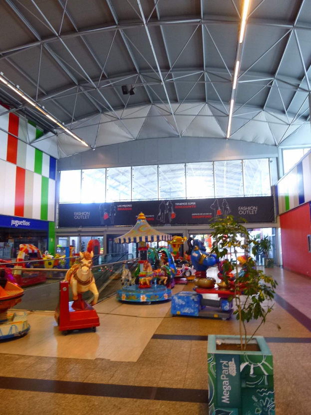 Großer Indoor-Vergnügungsbereich in einem Einkaufszentrum mit verschiedenen Fahrgeschäften, einem Topf mit einer Pflanze, verstreuten Spielzeugen, Informationsschildern, Deckenleuchten und Metallstützen.