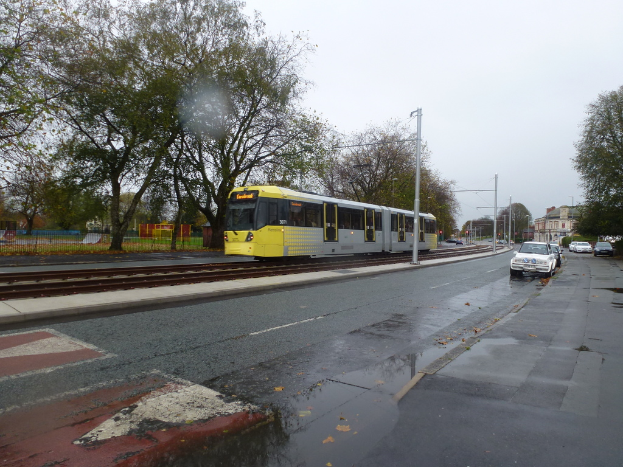 Eine belebte Straßenszene mit Fahrzeugen, Gebäuden, Pfählen, einer Bahnschiene mit einem Zug, Bäumen und einem Zaun unter einem klaren Himmel.