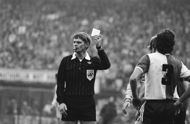 Schwarzes und weiße Bild eines Schiedsrichters mit einer roten Karte vor einer Menge von Fussballspielern, mit einem Zaun im Hintergrund.